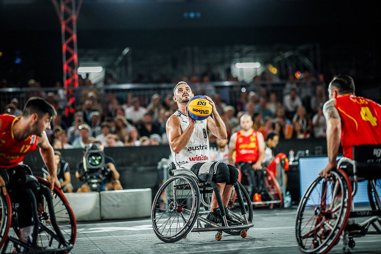 Der Rollstuhl-Basketballer Mehmet Hayirli hat ein weißes Trikot an und wirft aus dem Rollstuhl den Ball in Richtung Korb. Hinter ihm sieht man volle Zuseherreihen. 