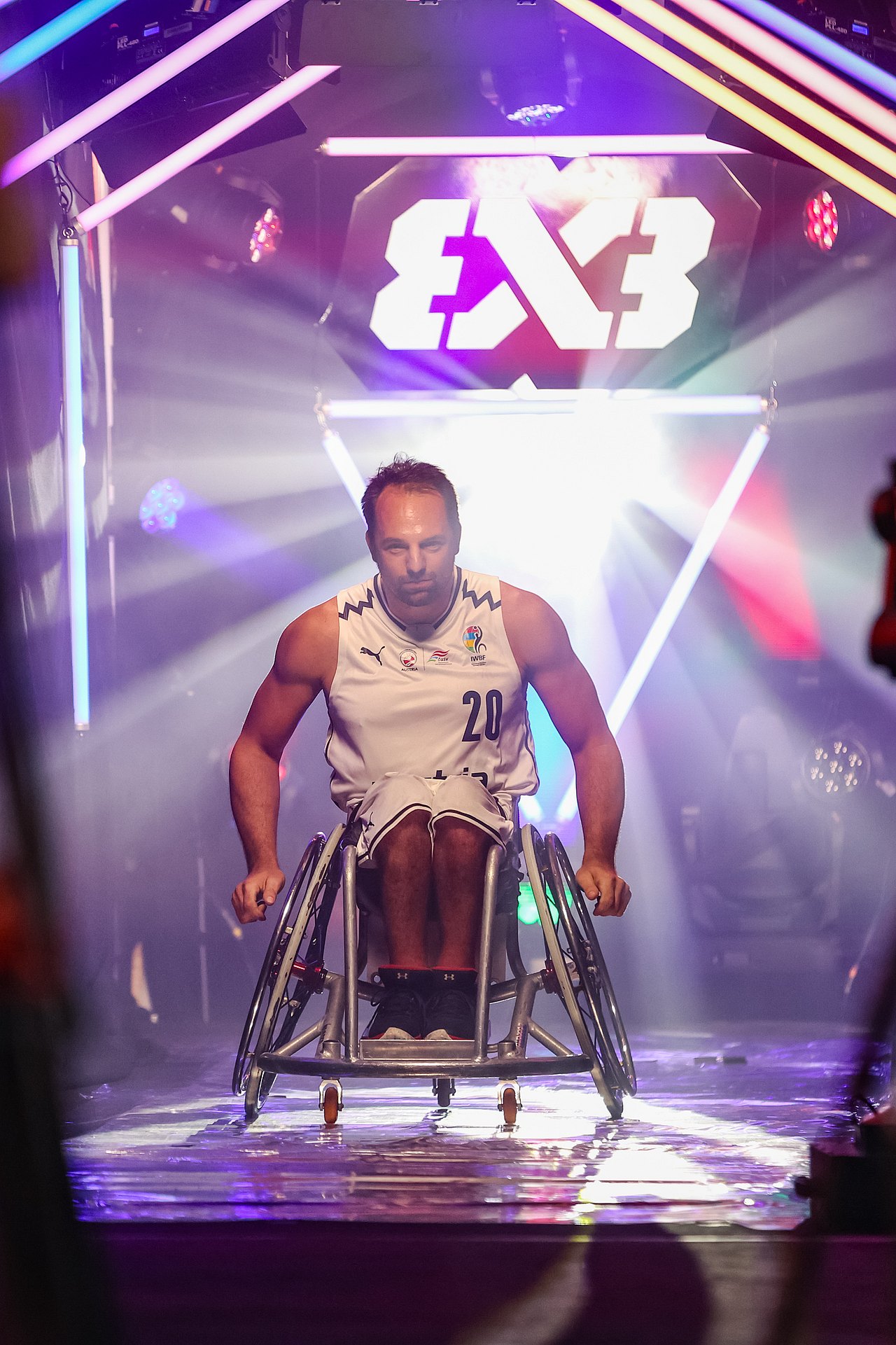 Matthias Wastian befindet sich im Spielertunnel bei der 3x3 EM in Wien. Er kommt in seinem Rollstuhl herausgerollt und trägt ein weißes Trikot.Violette und weiße Scheinwerfer umgeben ihn.