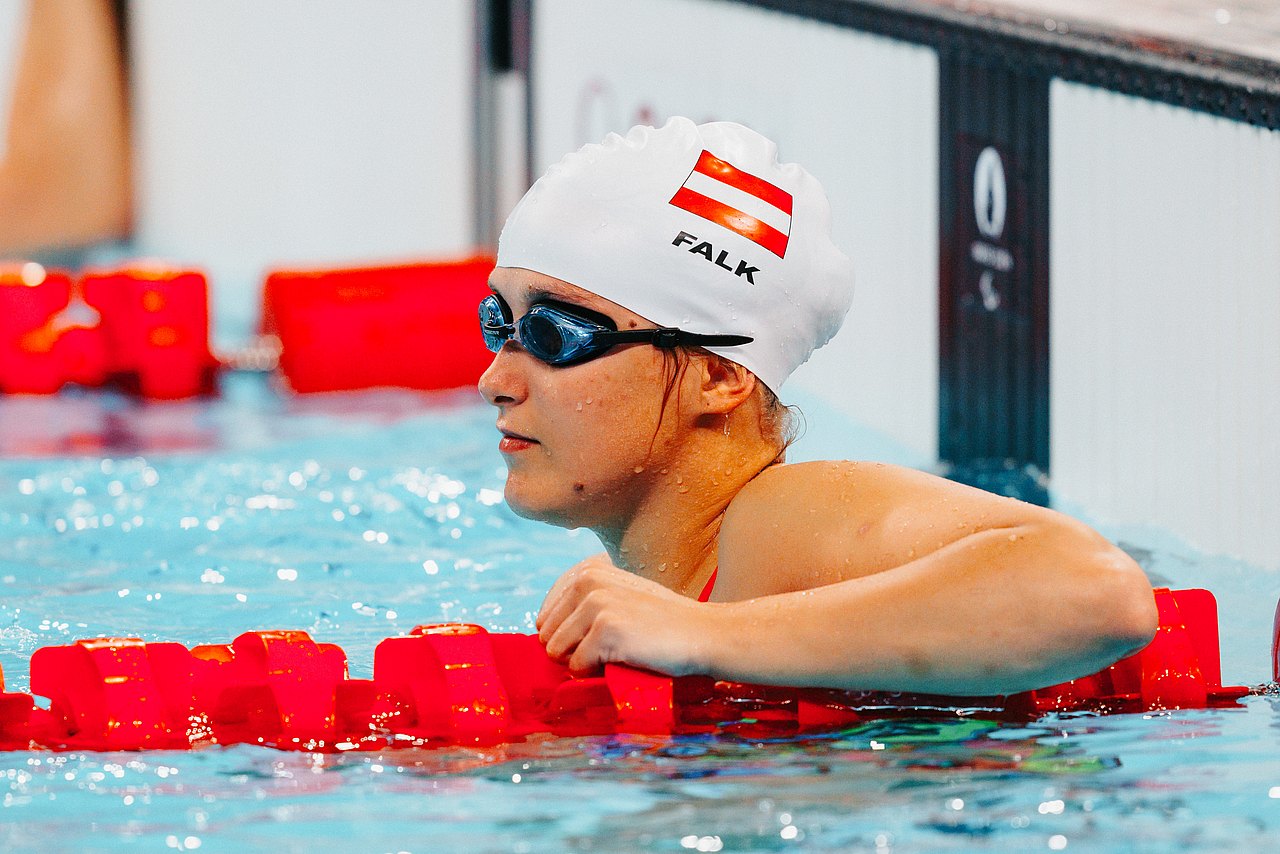 Janina Falk ist im Wasser und lehnt mit dem linken Arm an der roten Schwimmbahnbegrenzung. Sie trägt eine blaue Schwimmbrille und eine weiße Badehaube mit der österreichischen Flagge darauf. 