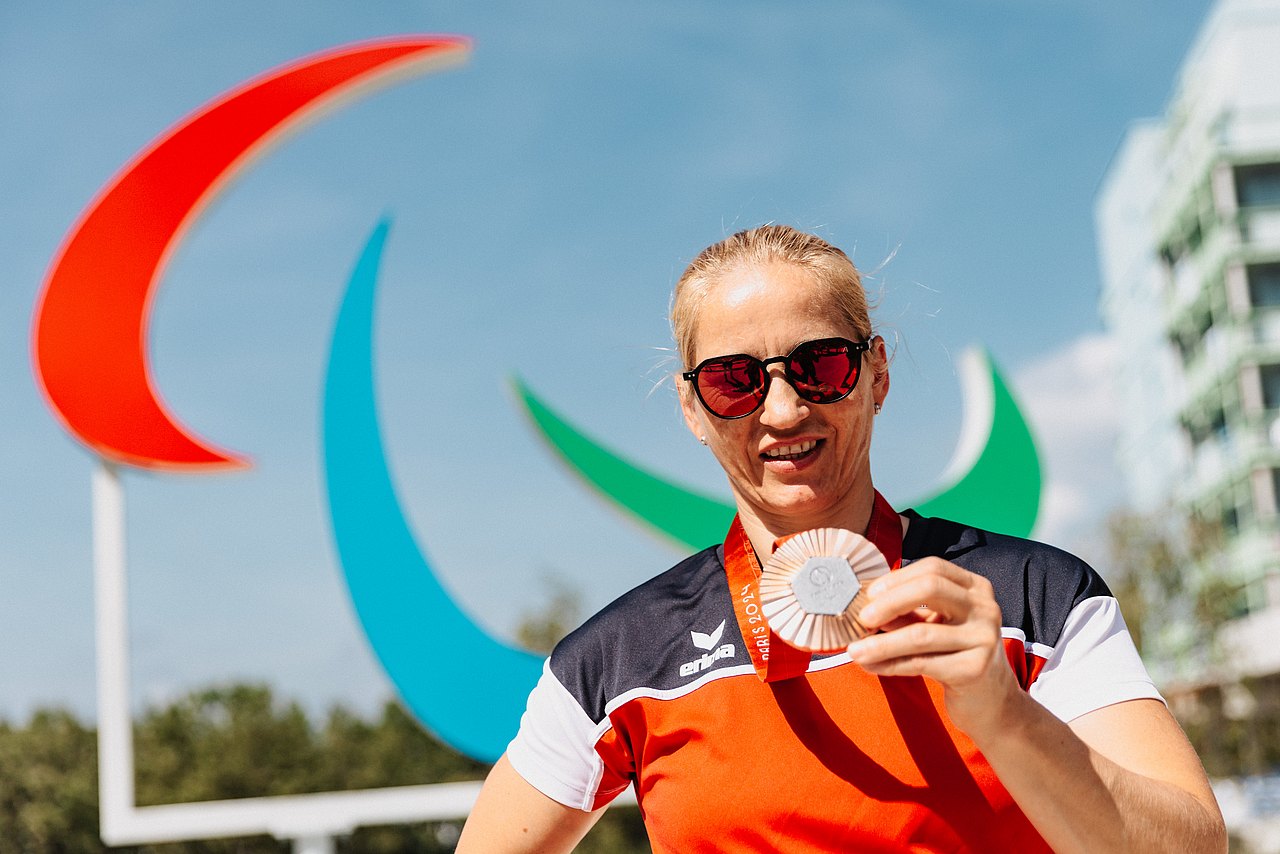 Natalija Eder hält ihre Bronzemedaille in der linken Hand. Im Hintergrund sieht man das Paralympische Zeichen. 