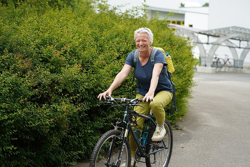 Birgit Offterdinger sitzt auf einem Fahrrad und lacht in die Kamera. Sie hat kurze blonde Haare, ein blaues Shirt und eine grüne Hose an. 