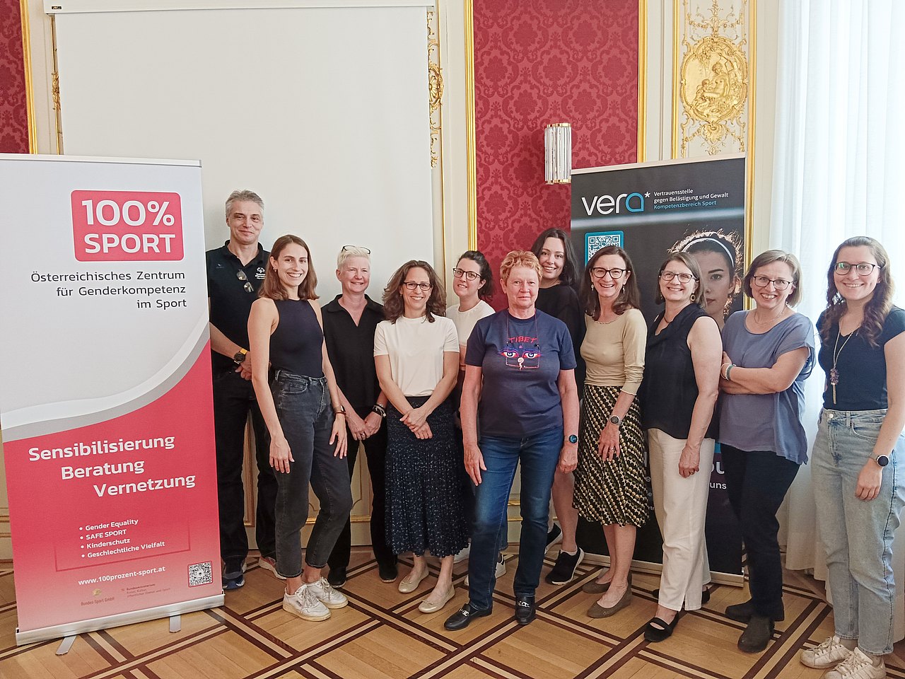 Die Teilnehmerinnen und Teilnehmer des Workshops stehen in einem prunkvollen Raum mit roten Wänden und lachen für ein Gruppenfoto in die Kamera. 