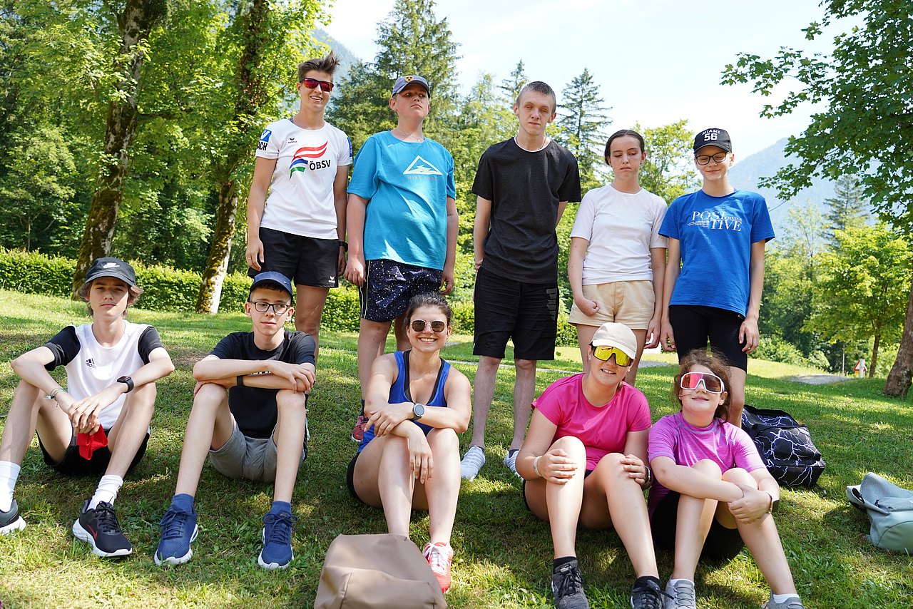 Kinder und Jugendliche sitzen und stehen auf einer Wiese. Sie tragen bunte T-Shirts, teilweise Sonnenbrillen und im Hintergrund befinden sich Bäume. Eine Person ist vom ÖBSV und trägt ein weißes T-Shirt mit dem ÖBSV-Logo.