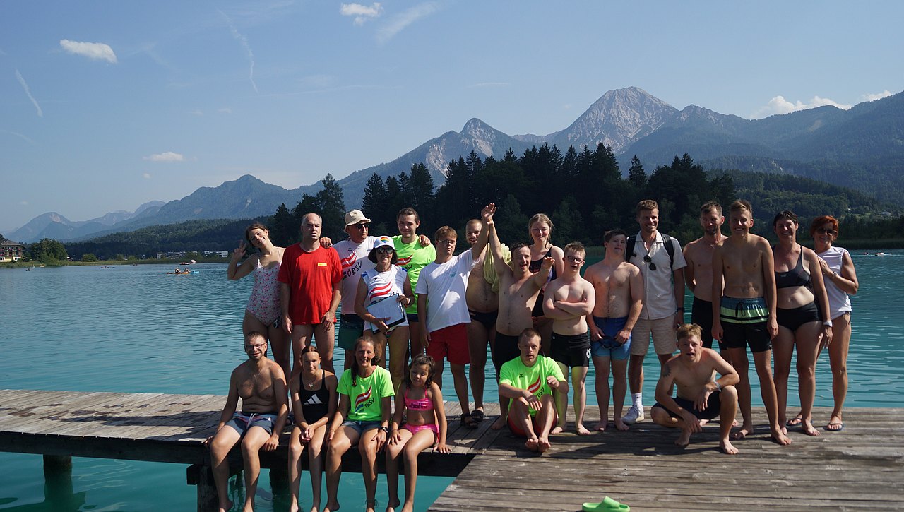 Die Teilnehmenden der Sportwoche stehen und sitzen auf einem Steg am See. Hinter ihnen sieht man ein schönes Bergpanorama. 