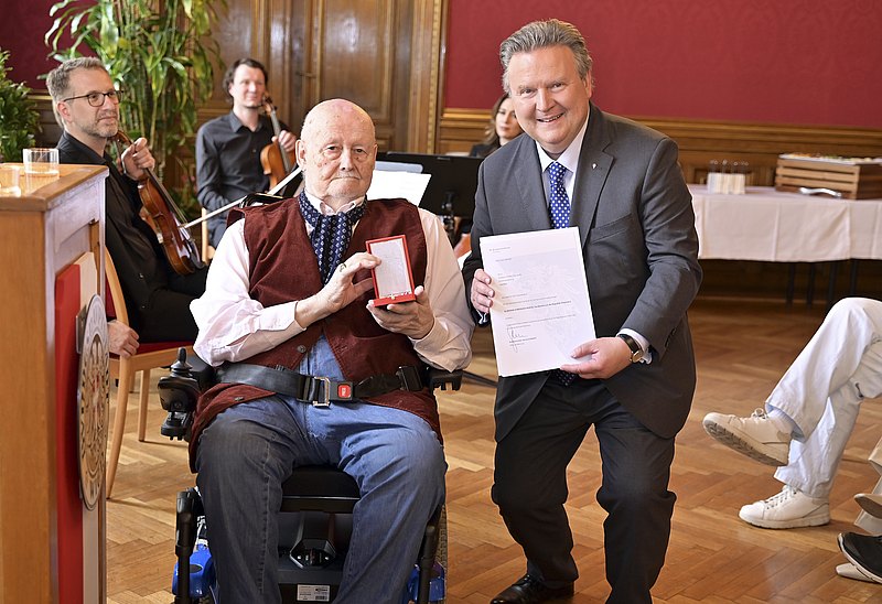 Franz Karl sitz im Rollstuhl. Er hält eine Schatulle mit dem Verdienstkreuz in die Kamera. Neben ihm steht leicht in der Hocke Wiens Bürgermeister Michael Ludwig, der der Kamera eine Urkunde präsentiert. 