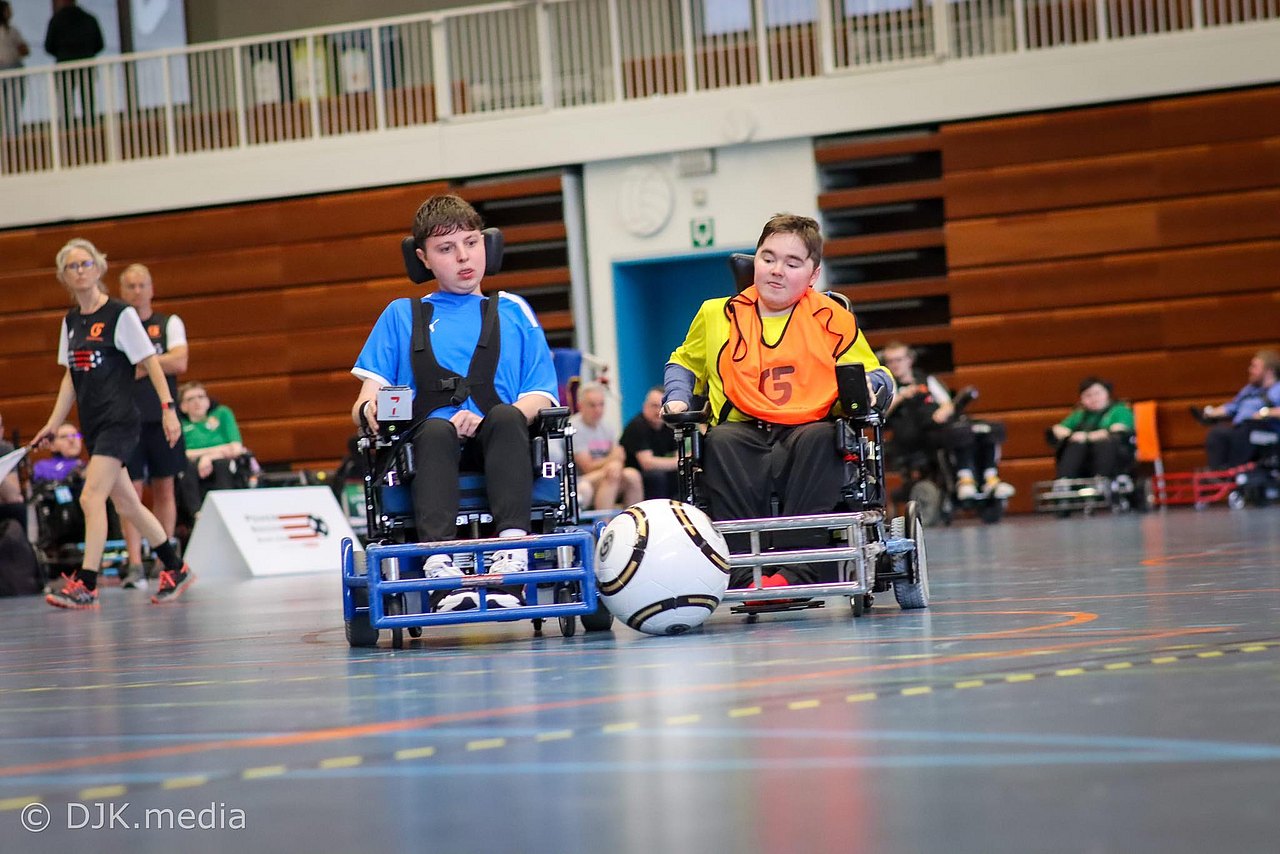 Zwei Spieler im E-Rolli liefern sich ein Duell um den Ball. Dieser ist größer als ein normaler Fußball. Sie spielen in einer Halle, der Ball befindet sich zwischen den E-Rollis. 