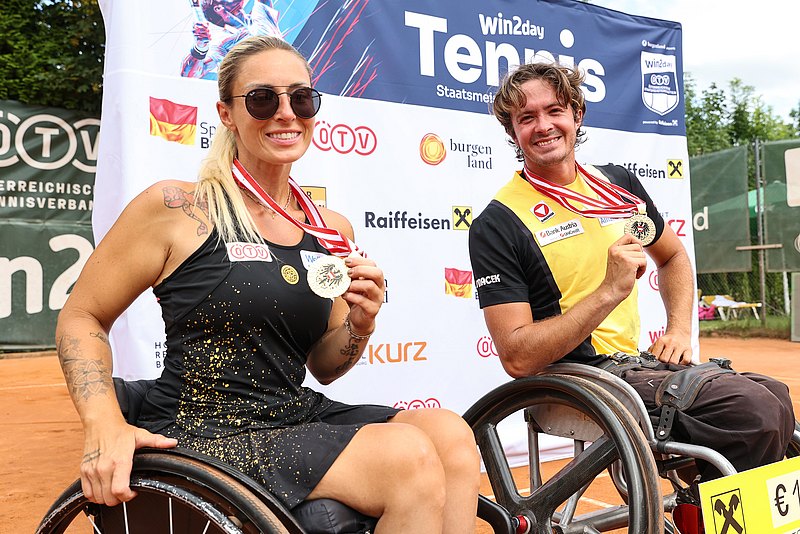 Die Rollstuhl-Tennisspieler Tina Pesendorfer und Nico Langmann stehen in ihren Rollstühlen vor einer Sponsorenwand. Beide halten ihre Goldmedaille in die Kamera. 