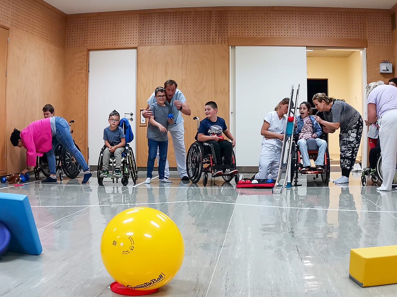 Drei Kinder im Rollstuhl und ein Kind, das steht, spielen in einem Turnsaal Boccia. Dabei werfen sie die Kugeln auf größere Ziele, wie zum Beispiel einen gelben Ball aus Plastik. 