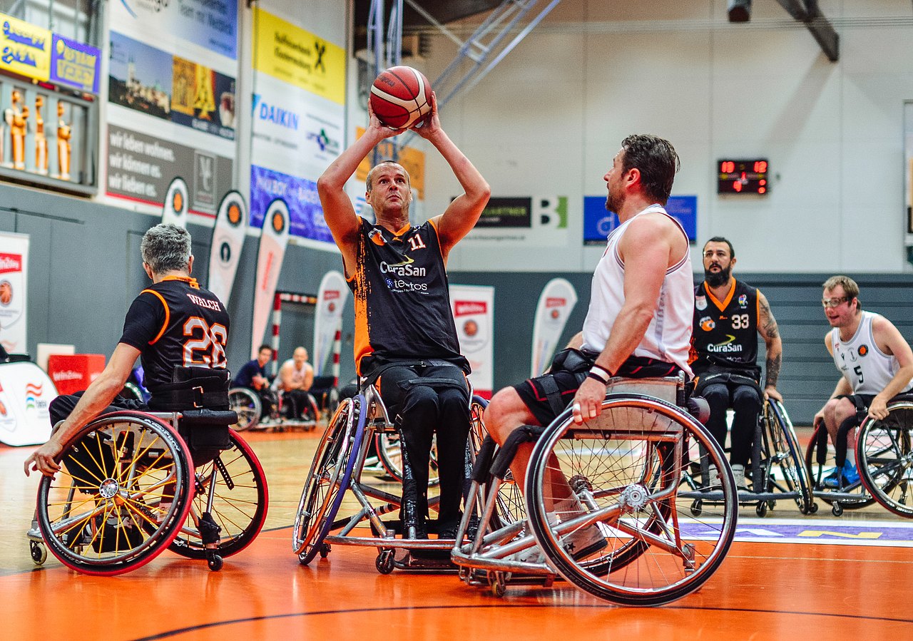 Ein Rollstuhl-Basketballer von den FlinkStones Graz hält den Ball mit beiden Händen hoch und setzt zum Wurf an. Er trägt ein schwarzes Trikot.