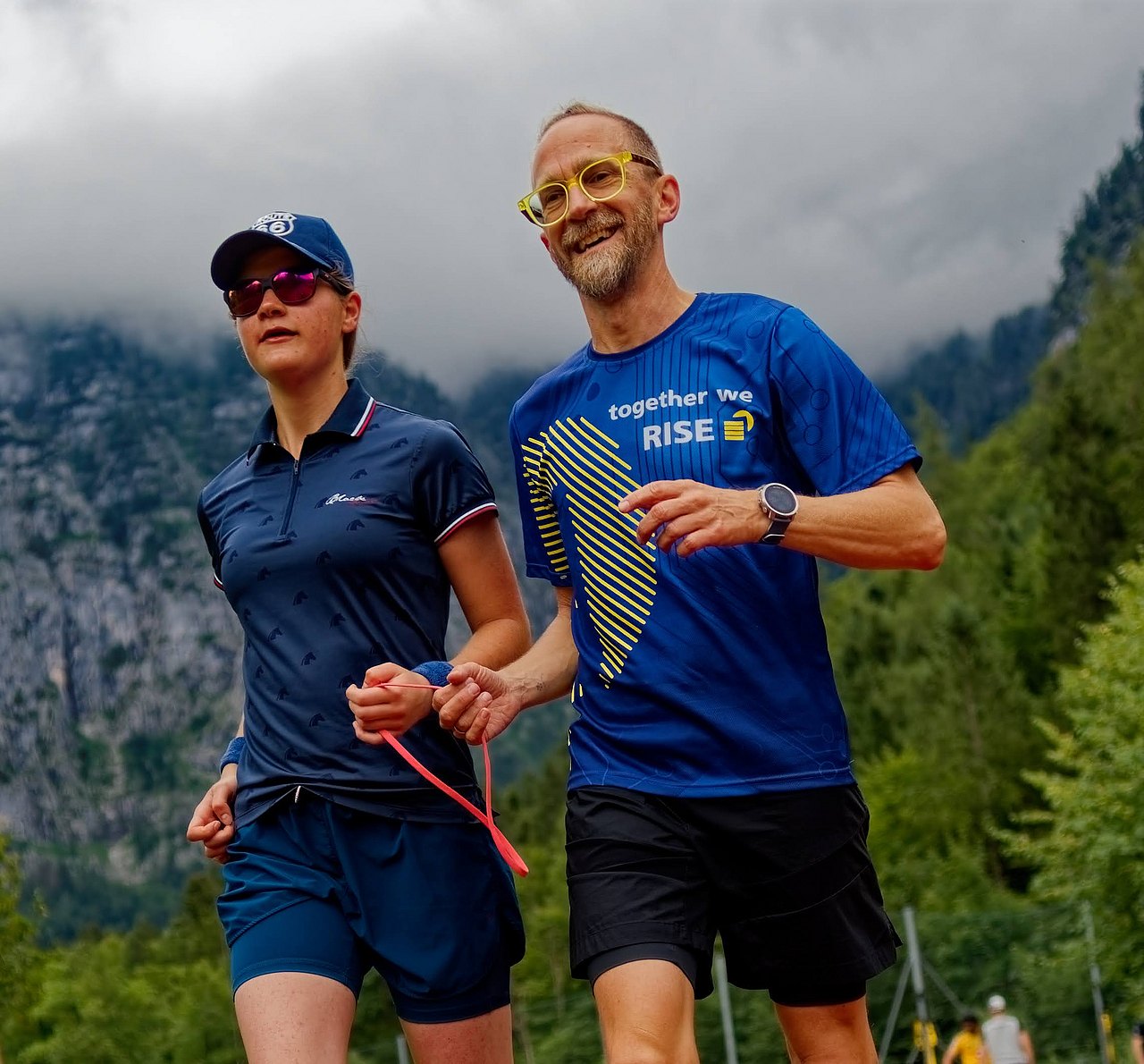 Die Blinden-Sportlerin Valentina Baier und ein Begleitsportler sind mit einem Band miteinander an den Händen verbunden und absolvieren eine Laufeinheit. Im Hintergrund befindet sich ein gewaltiges Bergmassiv im Nebel.