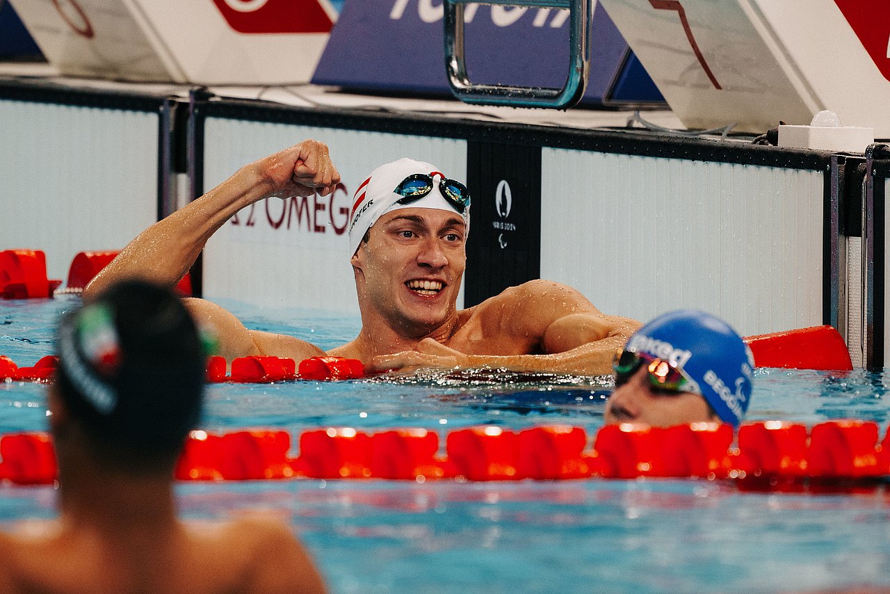 Andreas Ernhofer spannt nach Ende des Rennens seinen rechten Bizeps an. Er hat eine weiße Badekappe auf und lehnt auf der roten Schwimmbahnabtrennung. 