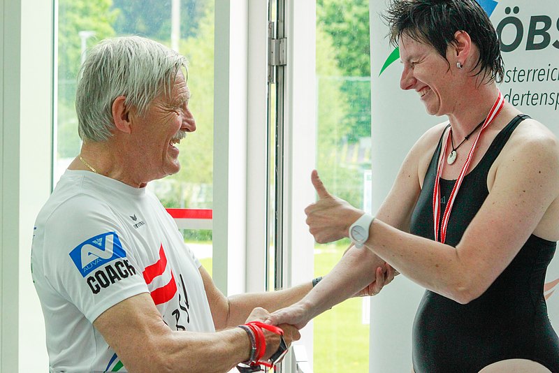 Werner Steindl schüttelt bei der Medaillenvergabe die Hand einer Athletin, die mit der anderen Hand den Daumen hochstreckt. 