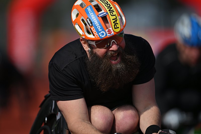 [Translate to Deutsch (Leichte Sprache):] Thomas Geierspichler sitzt in seinem Renn-Rollstuhl. Er hat eine Sonnenbrille und einen bunten Helm mit Sponsorenlogos auf. 