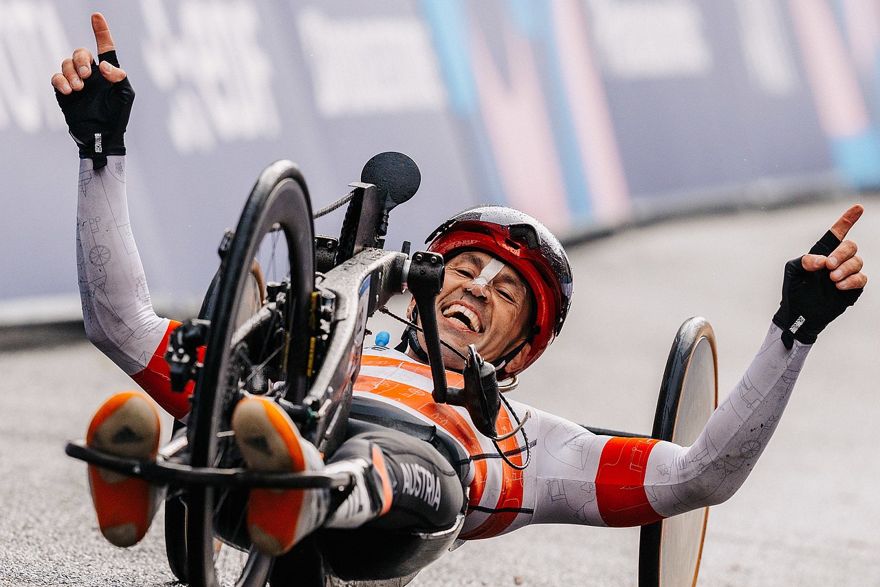 Thomas Frühwirth jubelt im Ziel liegend in seinem Handbike. Er streckt beide Arme in die Luft und hat ein breites Lächeln im Gesicht. Er trägt einen Helm und seinen österreichischen Rennanzug. 