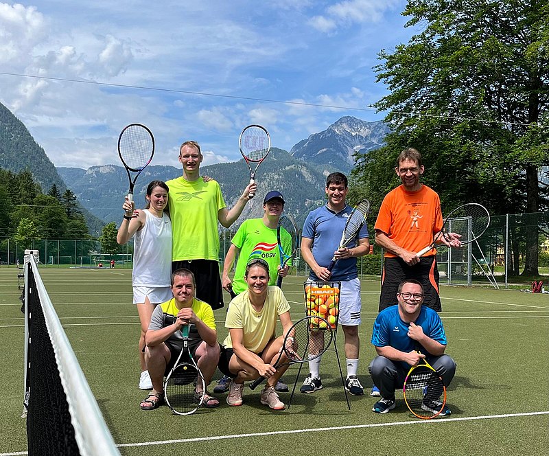 [Translate to Deutsch (Leichte Sprache):] Die Tennisgruppe steht für ein Gruppenfoto auf dem grünen Kunstrasen-Tennisplatz. Dahinter sieht man die schöne Gebirgslandschaft Oberösterreichs. 