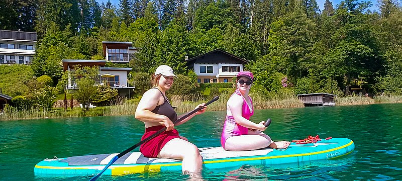 Zwei Sportwochen-Teilnehmerinnen sitzen im See auf einem Surf-Board und haben jeweils ein Padel in der Hand. Beide lachen in die Kamera. 