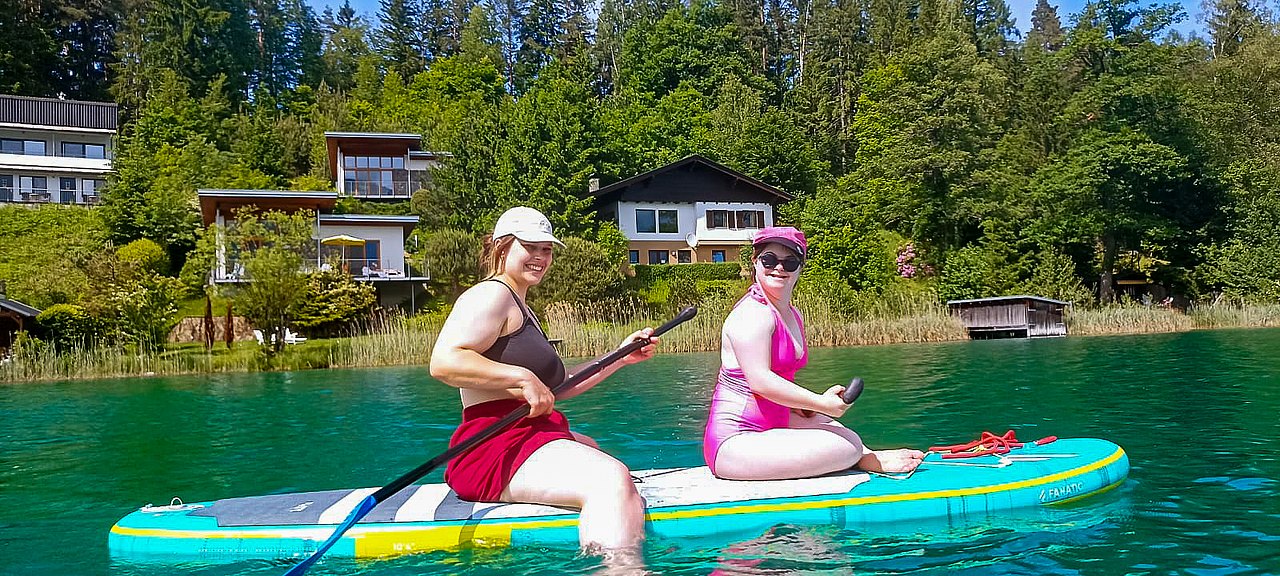 Zwei Sportwochen-Teilnehmerinnen sitzen im See auf einem Surf-Board und haben jeweils ein Padel in der Hand. Beide lachen in die Kamera. 