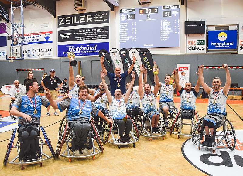 Die Rollstuhl-Basketball-Mannschaft der Sitting Bulls feiert in der Sporthalle. Ein Spieler stemmt den Pokal in die Höhe, die anderen strecken die Fäuste gen Himmel. 