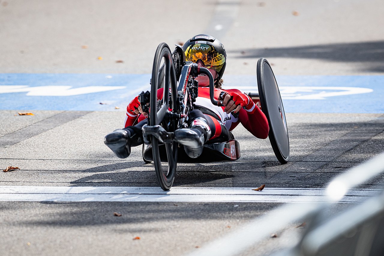 Svetlana Moshkovich liegt in ihrem Handbike und befindet sich im Straßenrennen. Sie trägt einen schwarzen Helm mit einer reflektierenden Schutzbrille und einen rot-weiß-schwarzen Rennanzug. 