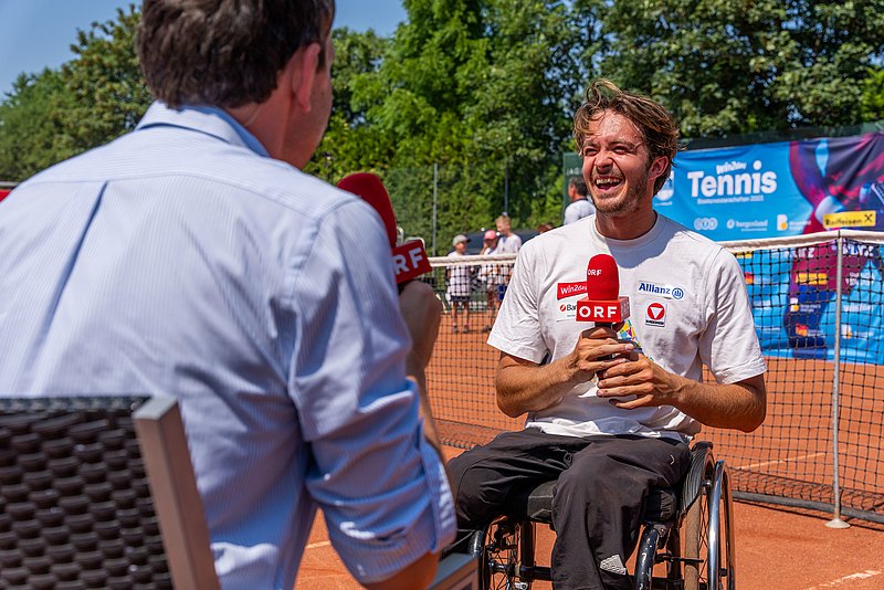 Rollstuhl-Tennisspieler Nico Langmann hält am Tennisplatz ein rotes ORF-Mikrophon in der Hand und lächelt den Interviewer an, der ihm gegenübersitzt. 