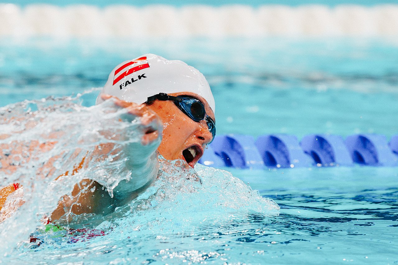 Die Schwimmerin Janina Falk im Wasser. Sie macht mit den Armen einen großen Schwung, hat eine Schwimmbrille auf und eine weiße Badekappe mit ihrem Nachnamen und der österreichischen Fahne auf.