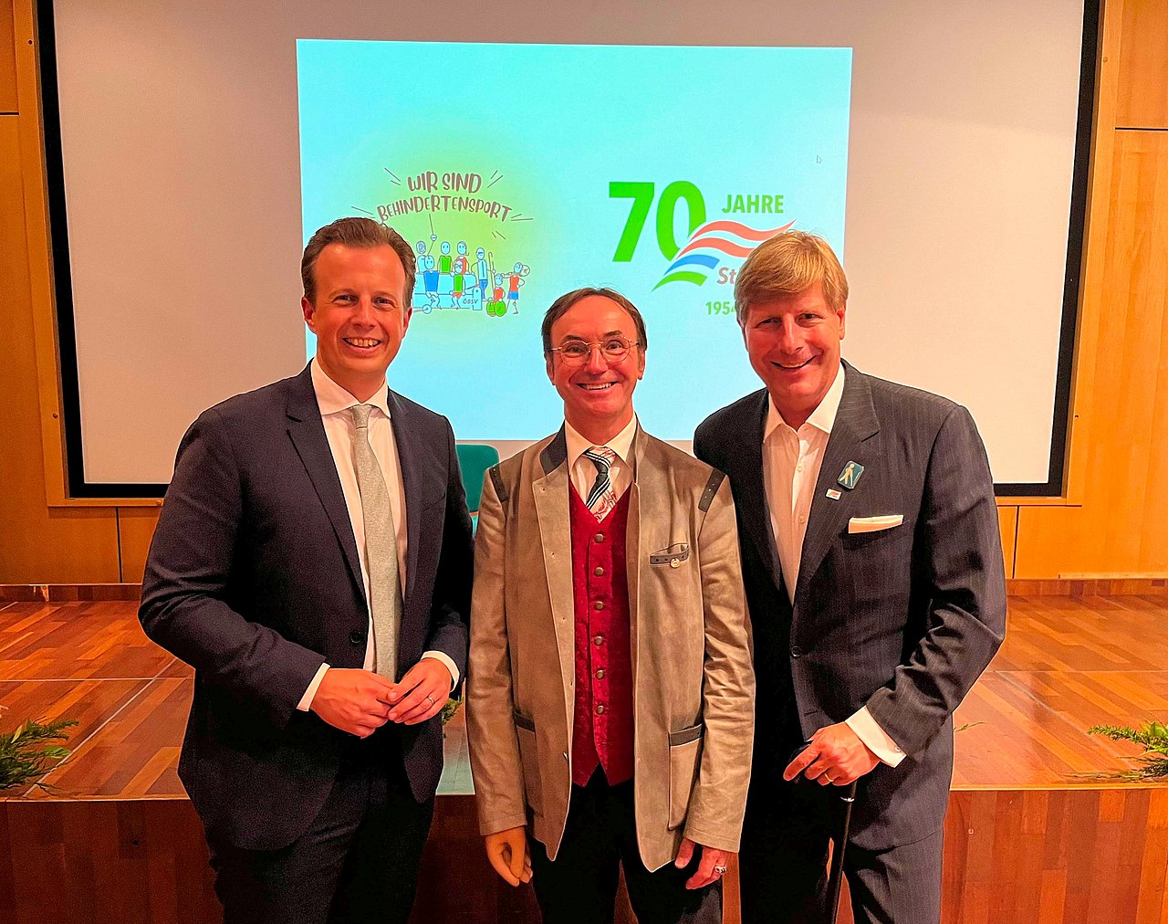 Karlheinz Kornhäusl, Herbert Rohrer und Julian Hadschieff stehen vor der Bühne. Sie sind festlich gekleidet und tragen einen Anzug. Im Hintergrund sieht man auf einer Leinwand 70 Jahre StBSV und das Logo Wir sind Behindertensport.