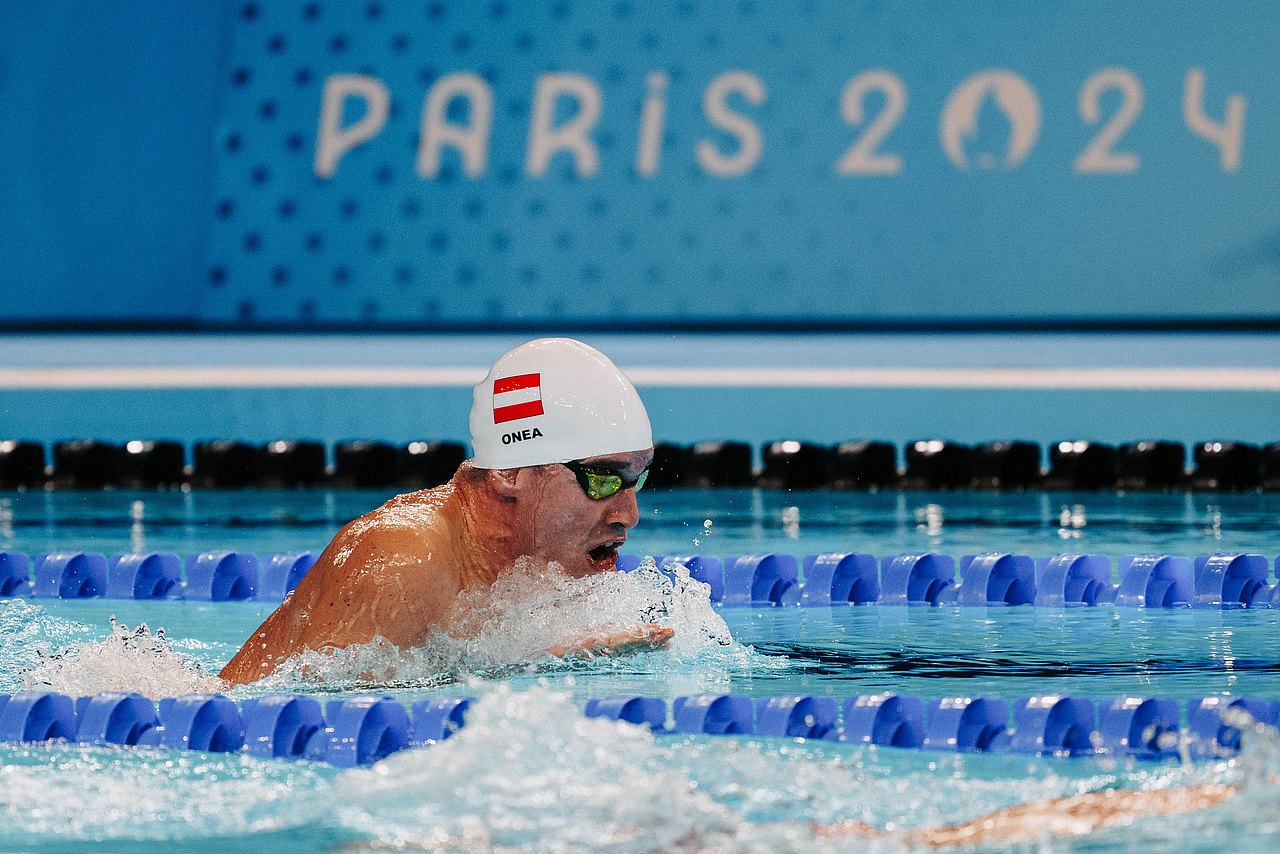 Der Schwimmer Andreas Onea im Becken. Sein linker Arm ist amputiert, er taucht mit dem rechten an. Er trägt eine weiße Badekappe mit seinem Nachnamen und der österreichischen Flagge drauf. Im Hintergrund steht "Paris 2024" geschrieben. 