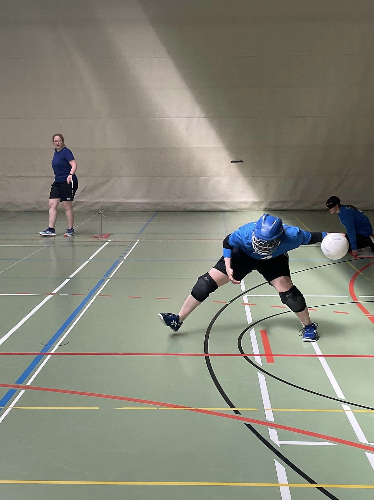 [Translate to Deutsch (Leichte Sprache):] Eine Spielerin holt weit mit der linken Hand aus, darin befindet sich ein weißer Ball. Sie trägt einen blauen Helm, passend zu ihrem blauen Trikot. 