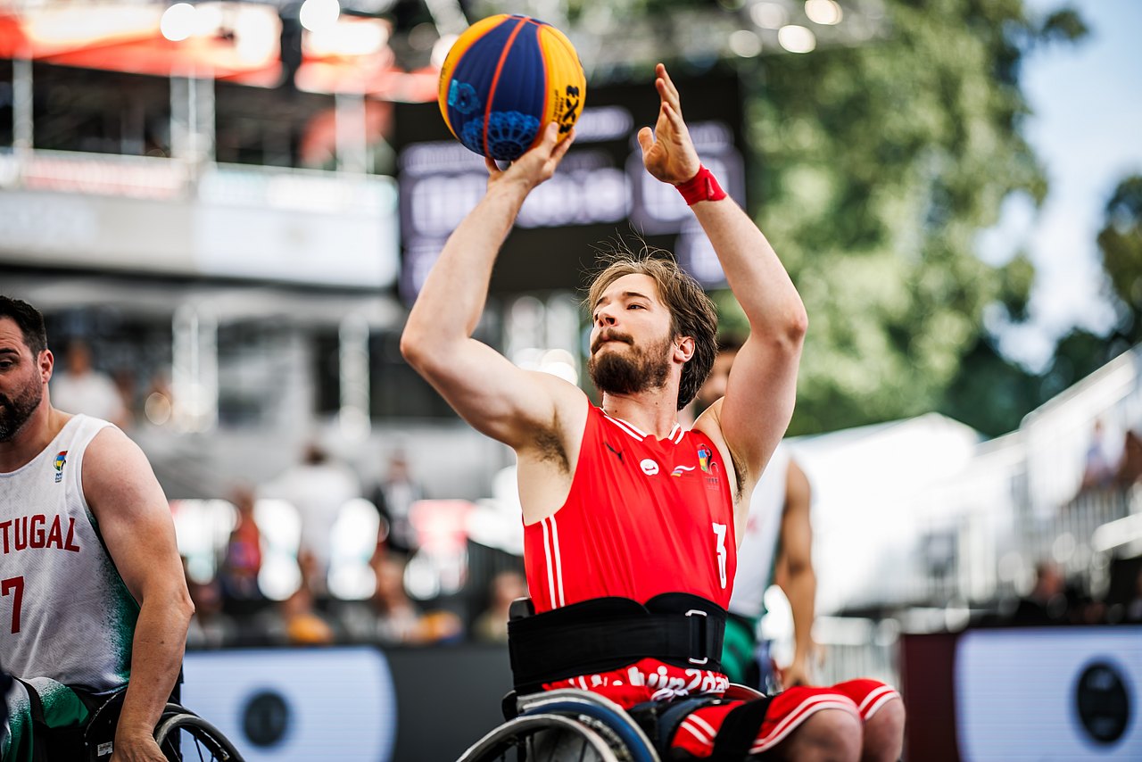 Der Rollstuhl-Basketballer Alex Suppan wirft aus seinem Rollstuhl den Ball in Richtung Korb. Er hat ein rotes Shirt an, hinter ihm sind die Zuseherränge zu sehen. 