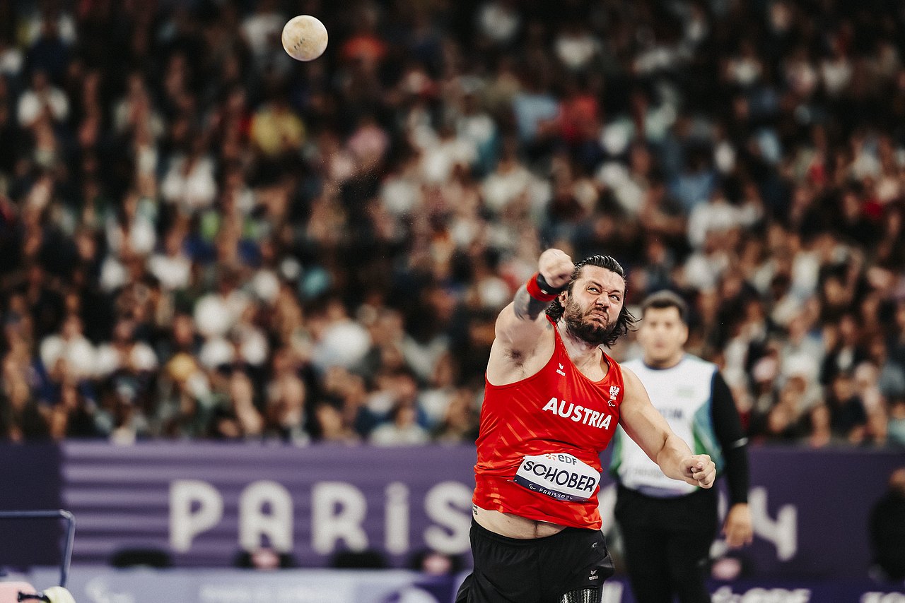 Georg Schober stößt mit seinem rechten Arm die Kugel hinaus. Er trägt eine schwarze Hose und ein rotes Shirt mit seinem Namen darauf. Im Hintergrund tobt das Publikum im Stade de France.