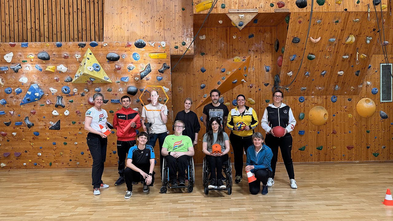 In einer Halle vor einer Kletterwand posieren 11 Personen für ein Gruppenfoto. 