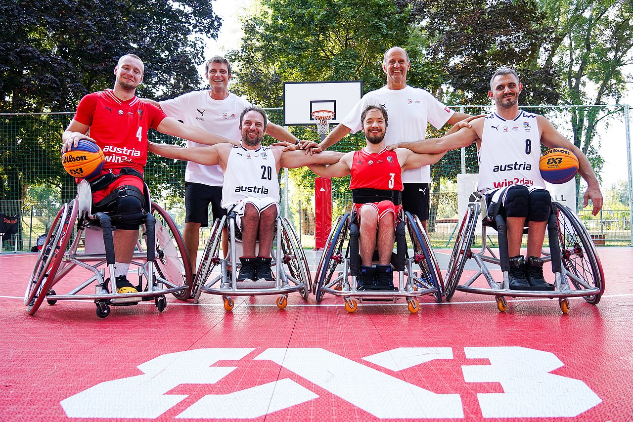 Das Rollstuhlbasketball-Team auf einem Gruppenfoto. Sie stehen auf einem roten Basketballplatz der mit dem Schriftzug 3x3 gekennzeichnet ist. Die Spieler und Betreuer haben die Arme über die Schultern des jeweils anderen gelegt. 