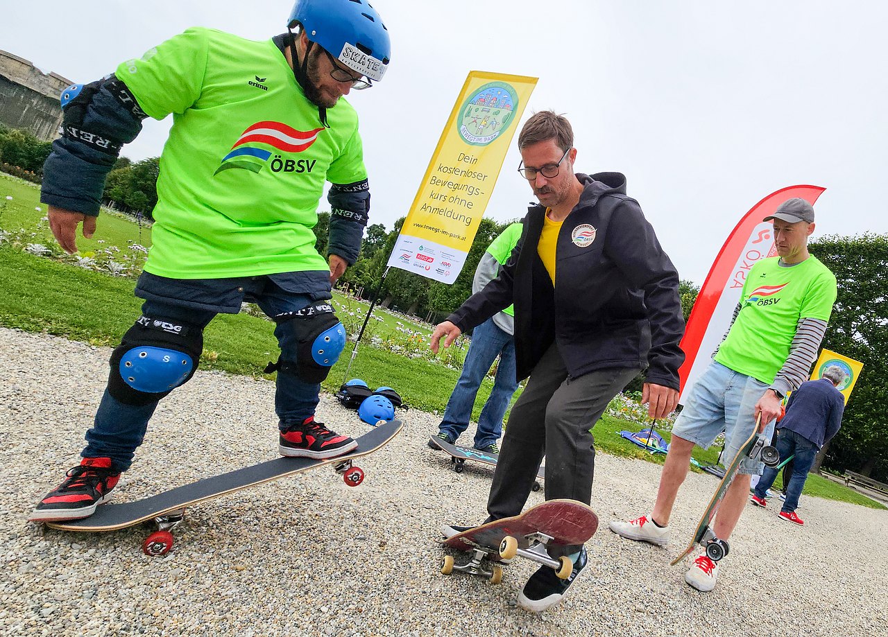 Coach Uwe Ballon zeigt einem Mann den Umgang mit dem Skateboard. Der HErr trägt ein grünes ÖBSV-Shirt, Knieschützer und einen Helm. Er steht am Skateboard und schaut dem Trainer bei der Erklärung der Übung zu.