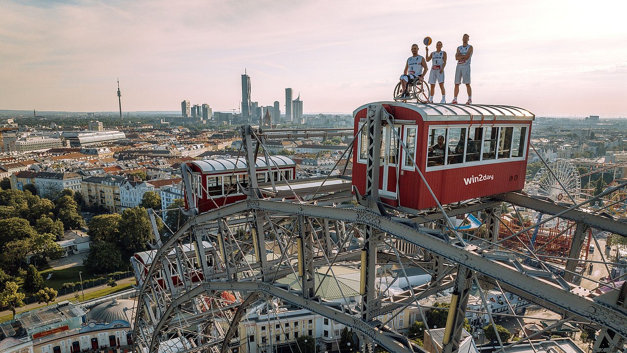 Die Basketballspieler Lisa Zderadicka, Mehmet Hayirli und Matthias Linortner stehen auf einem Wagon des Wiener Riesenrads in 70 Metern Höhe. Hayirli ist in seinem Rollstuhl an den Wagon geschnallt. Hinter ihnen ein toller Blick auf Wien.