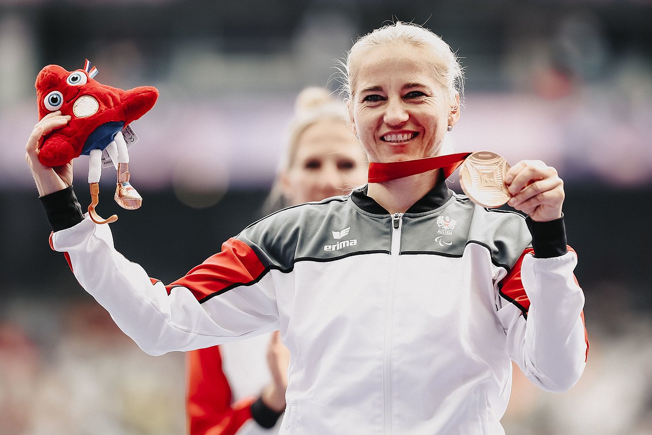 Die Speerwerferin Natalija Eder hält ihre Bronzemedaille in die Kamera. In der anderen Hand hält sie das rote Maskottchen. 