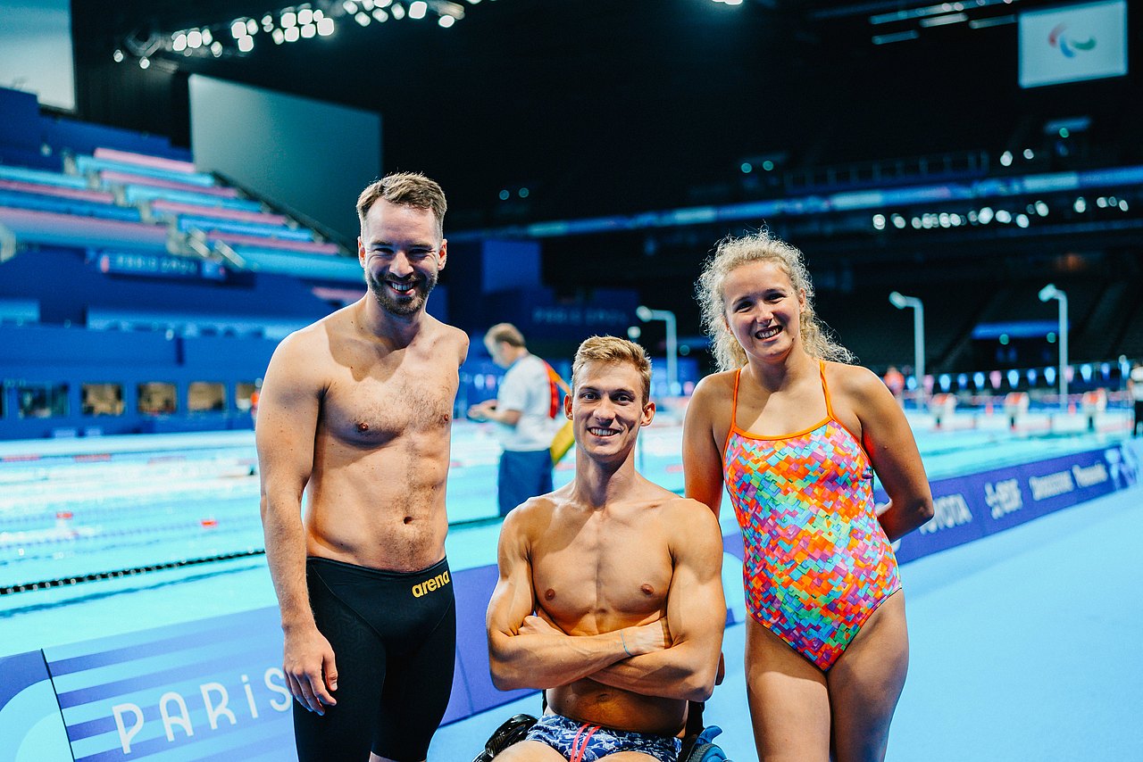 Andreas Onea, Andreas Ernhofer und Janina Falk stehen in der großen Schwimmhalle in Paris. hinter ihnen sieht man die Schwimmbecken und die Tribünen. 