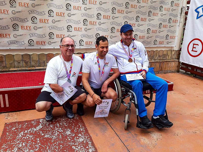Drei Männer, zwei davon knien einer sitzt im Rollstuhl, präsentieren ihre Medaillen und Urkunden. Hinter ihnen befindet sich eine Sponsorenwand, sie lachen an der Kamera vorbei. 