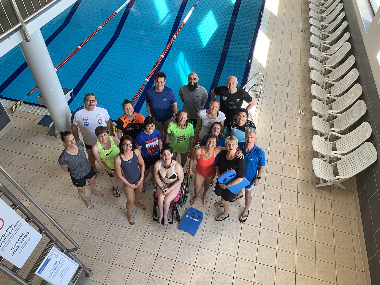 Die Kurs-Teilnehmer befinden sich am Beckenrand eines Indoor-Pools und blicken hinauf in die Kamera, die von einer Erhöhung fotografiert. 