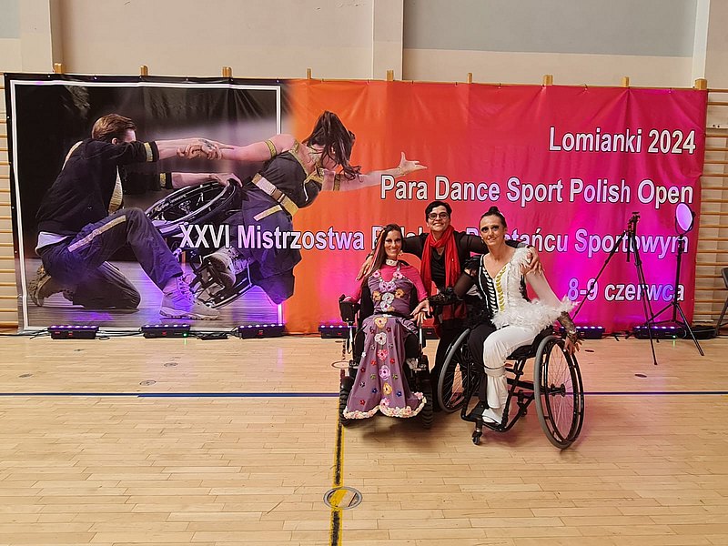 Die Tänzerinnen Eva-Maria Nussdorfer im E-Rolli und Sanja Vukasinovic im Rolli stehen gemeinsam mit ihrer Trainerin Kerstin Govekar vor einem bunten Banner der Veranstaltung. 