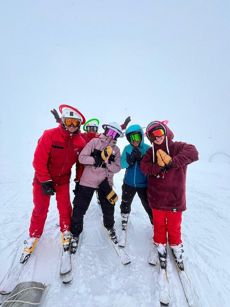 Fünf Personen stehen in voller Ski-Ausrüstung auf einer Piste und lachen in die Kamera. 