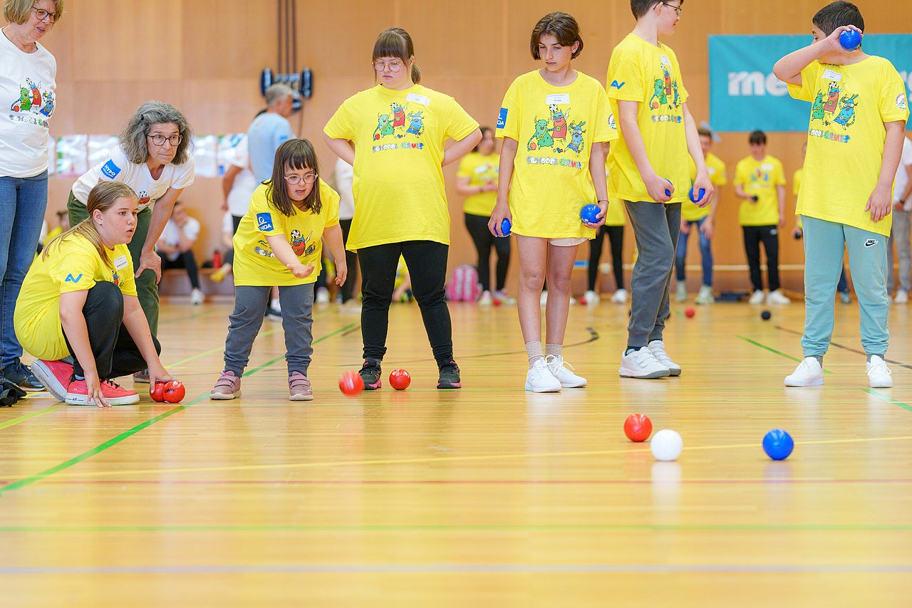 Ein Mädel mit Downsyndrom spielt eine rote Boccia-Kugel. Zwei Teamkolleginnen und eine Lehrerin schauen ihr dabei zu. Sie spielen in einer Sporthalle. 