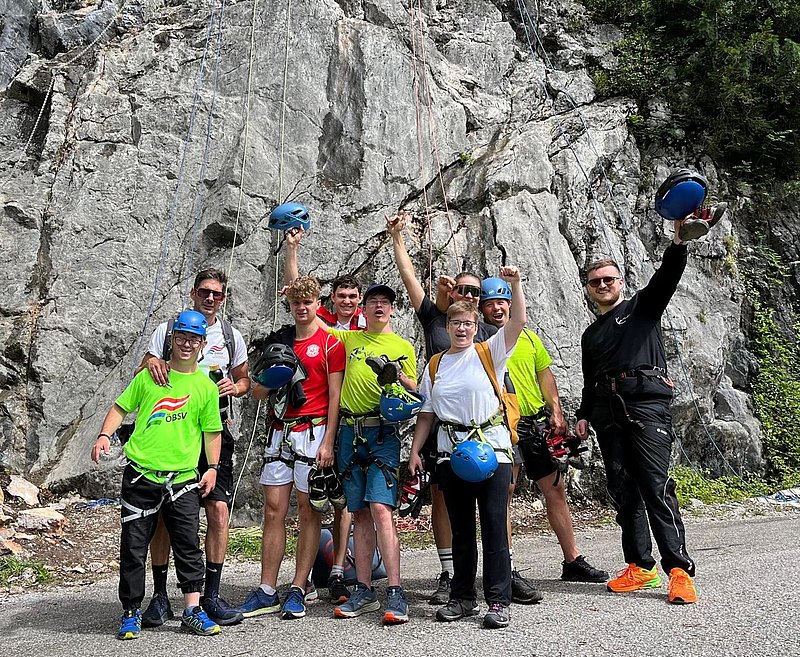 Die Sportgruppe steht vor einer Felswand. Alle haben Helme und Sicherungsgurte an. Sie lachen in die Kamera und strecken die Hände in die Höhe. 