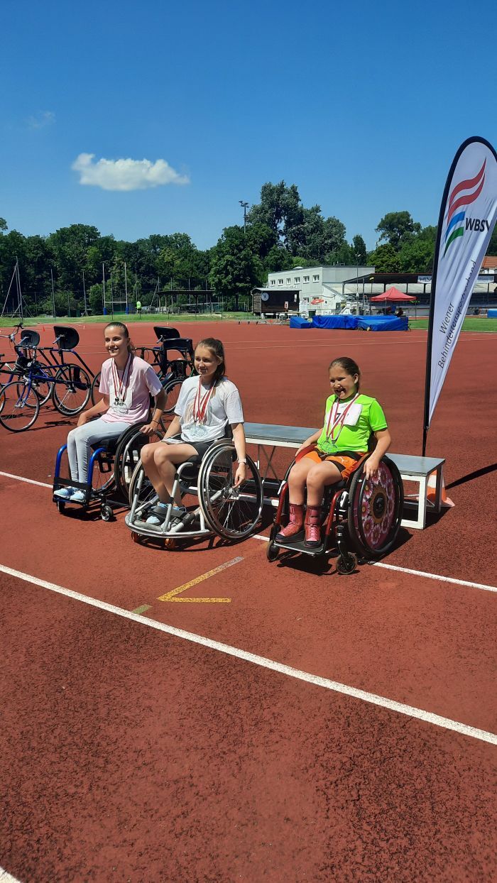 Drei Sportlerinnen im Rollstuhl stehen bei der Siegerehrung auf der roten Laufbahn den Leichtathletik-Zentrums und haben ihre Medaillen umgehängt. 