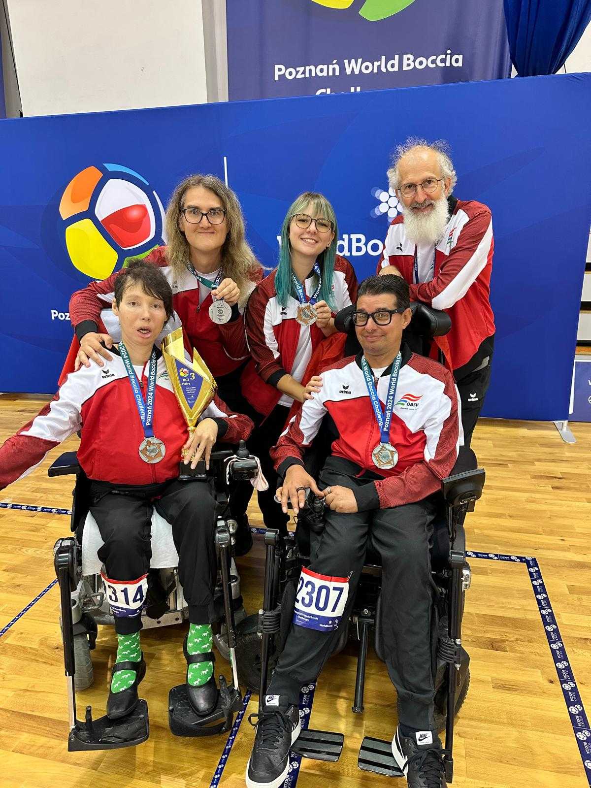 Die beiden Spielerinnen und Spieler Michael Kiefler und Steffi Proßegger sitzen in ihren E-Rollis mit der Bronzemedaille um ihren Hals. Proßegger hält einen goldenen Pokal in der linken Hand. Dahinter stehen ihre Betreuerinnen und ihr Coach. Alle lachen in die Kamera. 