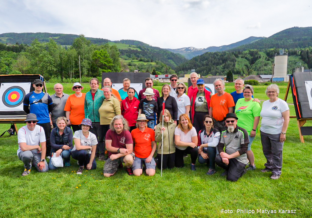 Die Teilnehmerinnen und Teilnehmer stehen und sitzen für ein Gruppenfoto auf einer Wiese. Hinter ihnen sind Zielscheiben für den Bogensport aufgebaut. 