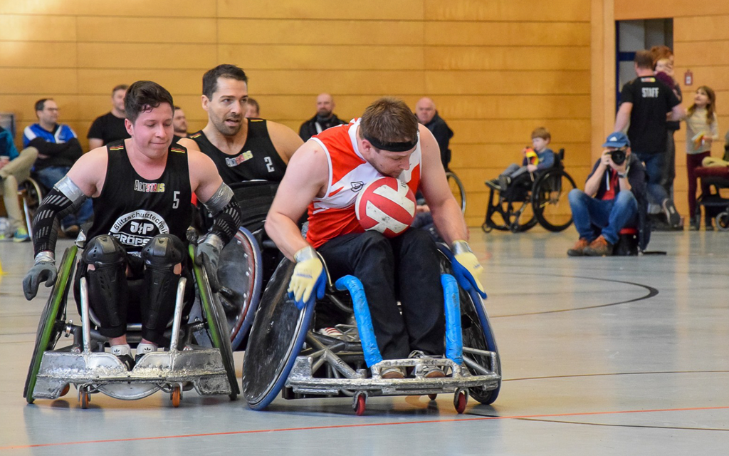 Ein Rollstuhl-Rugby-Spieler Österreichs hat sich den Ball in den Schoß gelegt und fährt in Richtung Torzone. Zwei Spieler Deutschland fahren hinterher. 