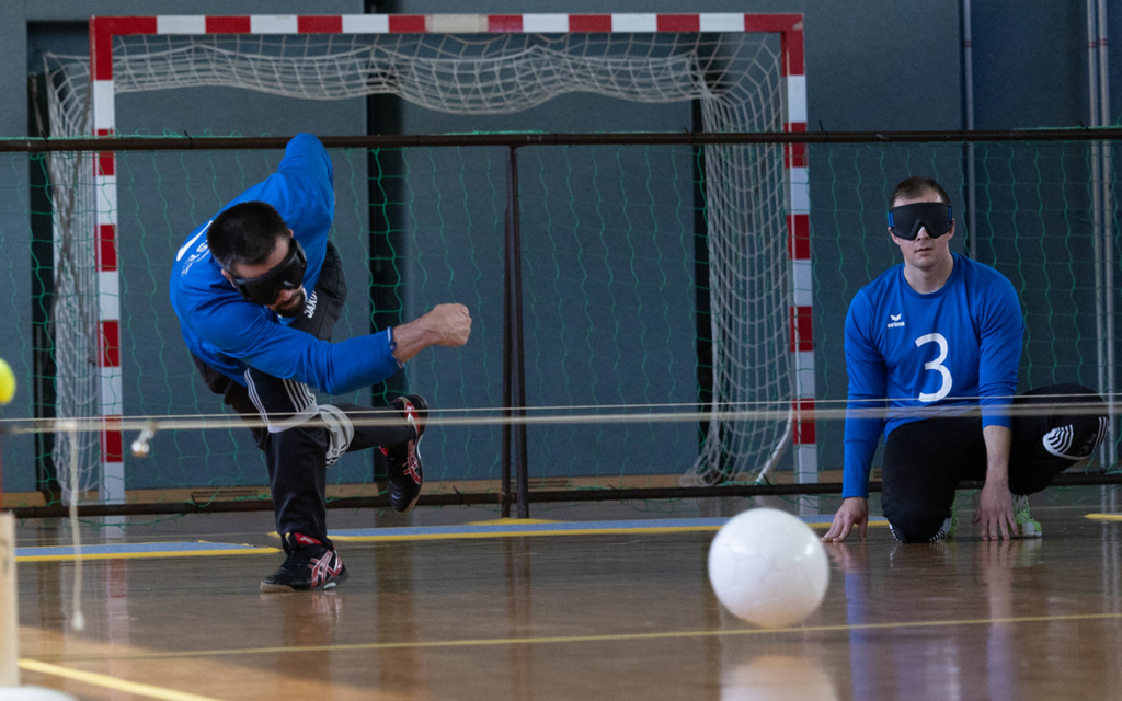 [Translate to Deutsch (Leichte Sprache):] Ein Torballspieler rollt den Ball mit viel Kraft den Boden entlang unter einer Schnur durch. Ein Mitspieler sitzt aufmerksam dahinter. 